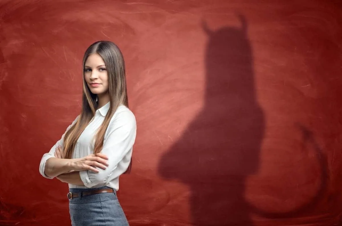 Woman with crossed arms in front of a red wall; her shadow, resembling an insurance adjuster, appears with horns and a tail.