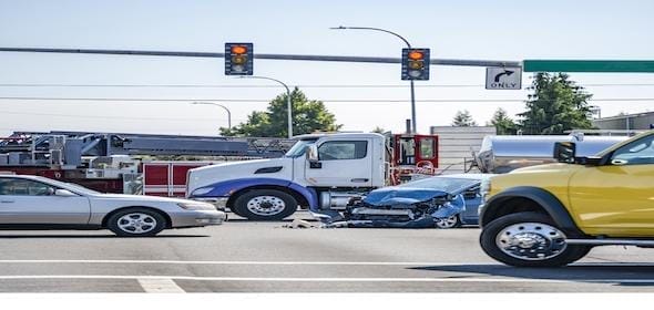 A traffic accident at an intersection involved a truck and a blue car with front damage, while a yellow vehicle passed by. Overhead, traffic lights blinked knowingly, as if aware of the potential injuries to any accident victim caught in such a situation.