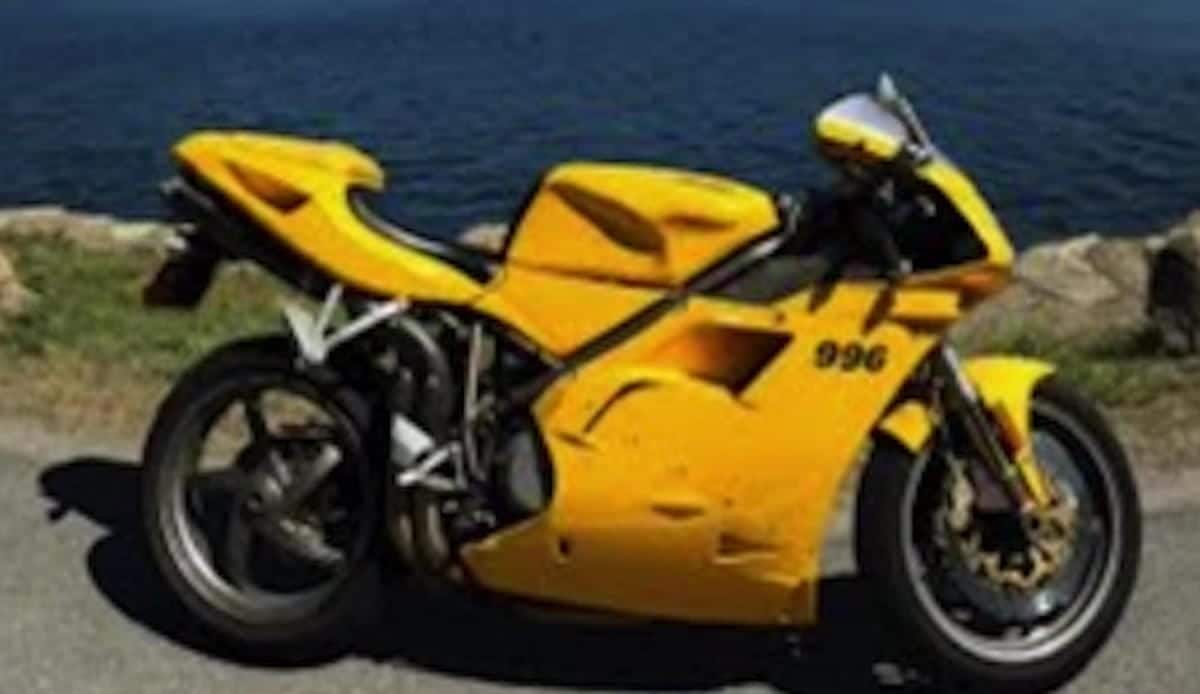 A yellow sport motorcycle with "996" on the side, parked beside a body of water, has a sleek motorcycle helmet resting on its seat.