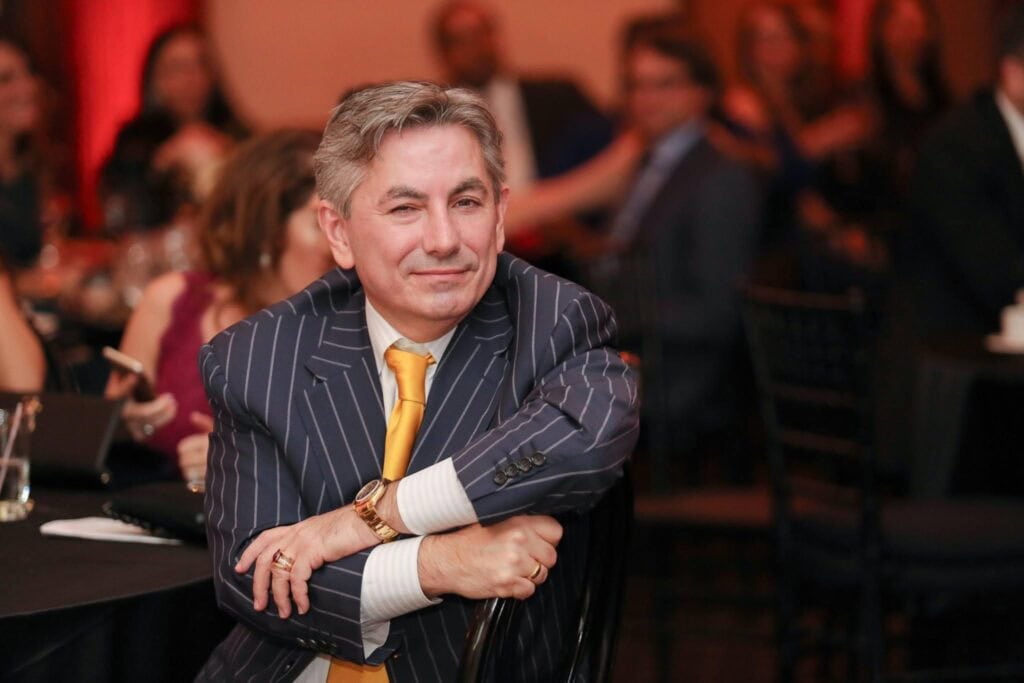 A man in a pinstripe suit and yellow tie sitting with his arms crossed, smiling, at an indoor event.