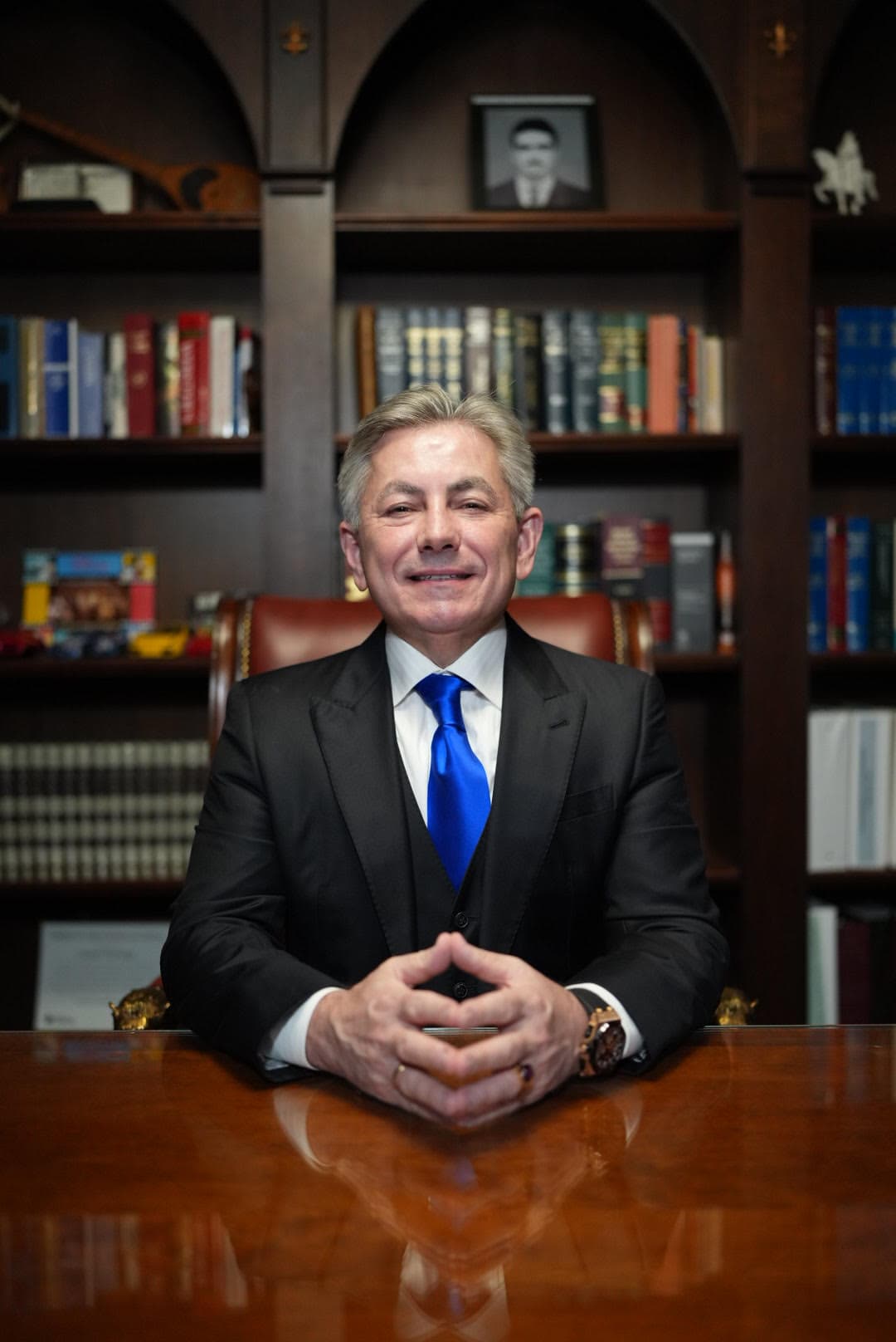A person in a suit sits at a wooden desk with hands clasped. Bookshelves filled with books and framed pictures are in the background.