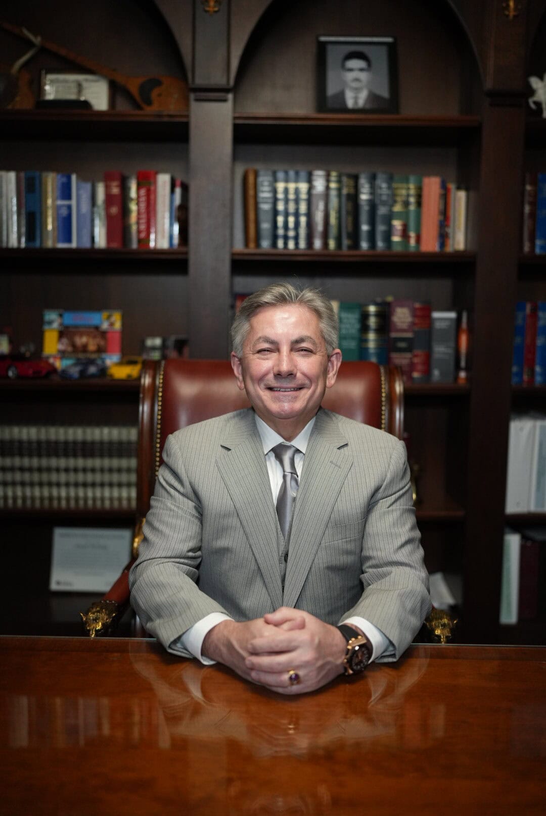 A man in a suit sits at a polished wooden desk, smiling, with bookshelves and a framed photo in the background.