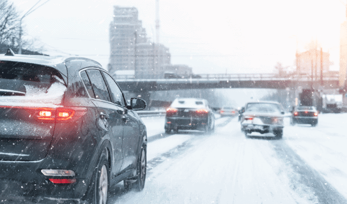 Cars navigate a snow-covered road in a Michigan city, the backdrop of falling snow and towering buildings highlighting the challenges of winter weather conditions.