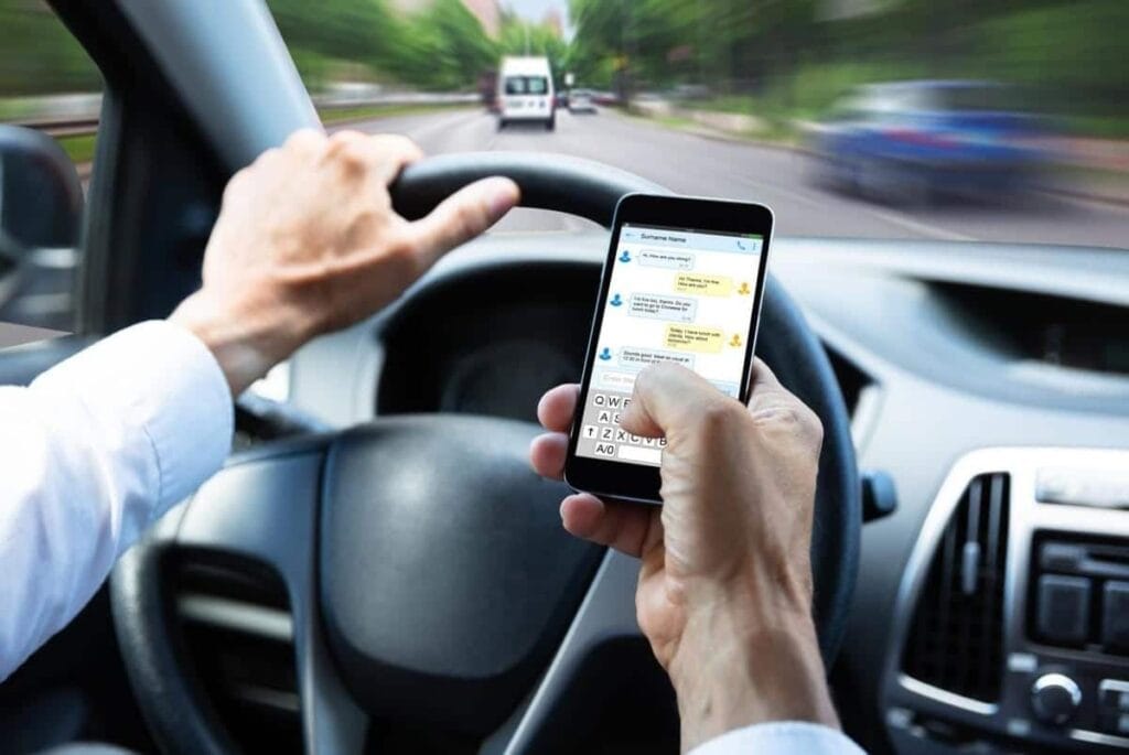 The driver, engaging in distracted driving, uses a smartphone to text while steering the moving car, with a blurred view of the road visible through the windshield.