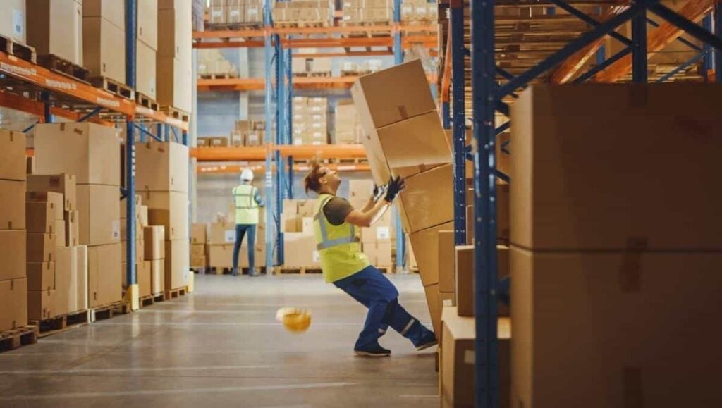A worker in safety gear tries to stabilize a tumbling stack of boxes in a warehouse aisle, highlighting the importance of avoiding slip and fall accidents.