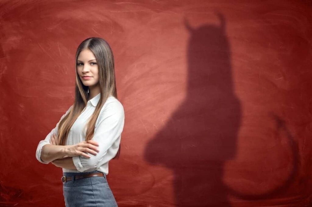 Woman with crossed arms in front of a red wall; her shadow, resembling an insurance adjuster, appears with horns and a tail.