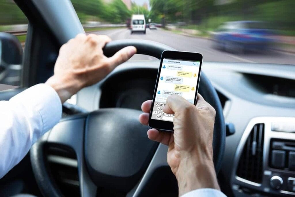A person is texting on a smartphone while driving a new car. The steering wheel is in the left hand, and the road is visible through the windshield, raising concerns for the U.S. DOT as it sets fuel economy standards for safer driving practices.