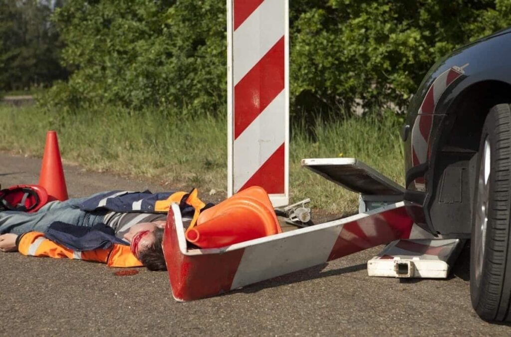 A person in a safety vest lies on the road with an overturned traffic barrier and cones near a car, suggesting a Michigan work-related car accident beside a grassy area and trees.