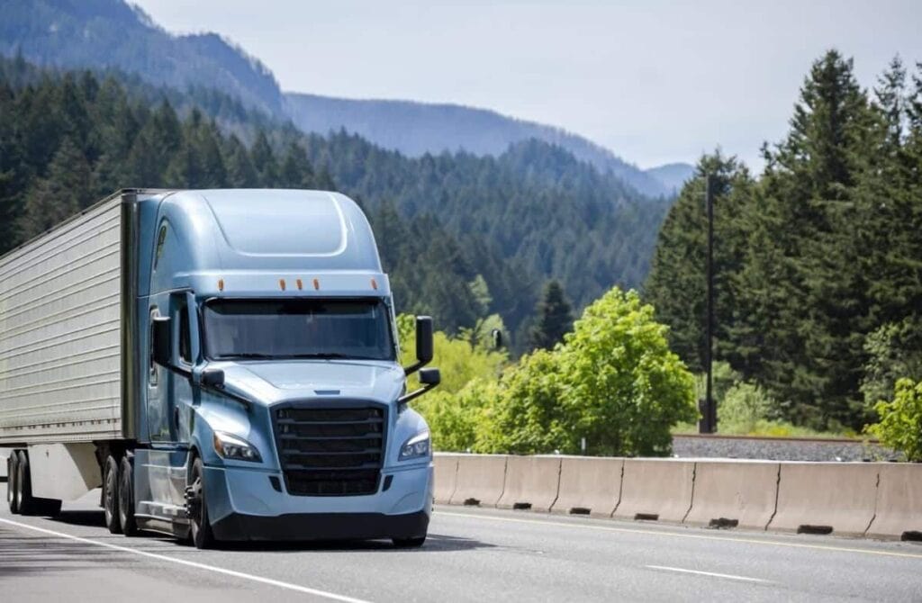 A blue semi-truck with a large trailer travels on a highway, surrounded by green trees and majestic mountains, maximizing its journey through nature's beauty.
