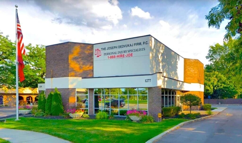 Exterior of The Joseph Dedvukaj Firm, P.C., a personal injury law office specializing in car accident liability, with a sign displaying contact information. An American flag and landscaped greenery are visible outside.