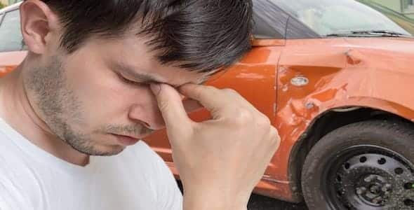 A man in a white shirt holds his face in frustration after a minor accident. Behind him, an orange car, damaged on the side, awaits an insurance claim.