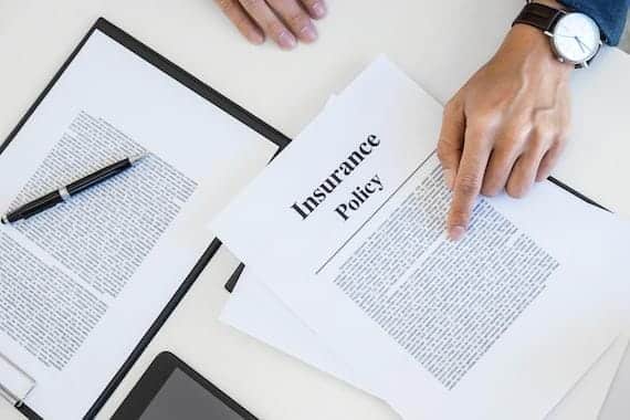 A person points to a printed insurance policy document on a table, offering insurance advice. Nearby, there's a clipboard with a pen and part of a digital device.