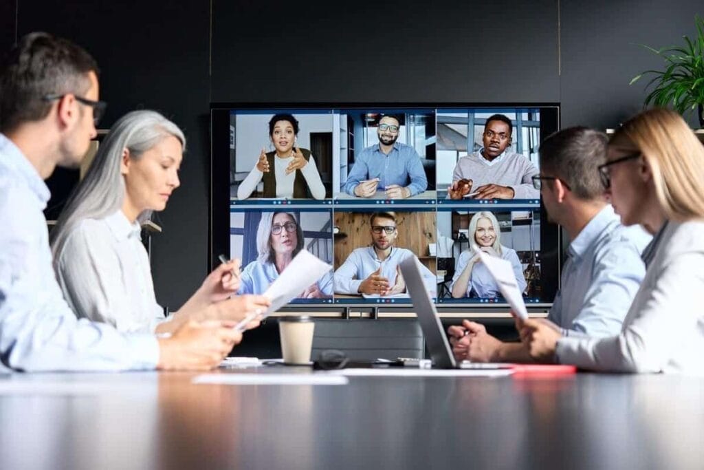 People in an office meeting room are working together, participating in a video conference using Zoom technology. They view several colleagues displayed on a large screen, enhancing collaboration and communication.