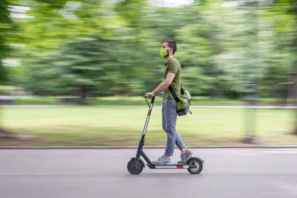 Wearing a green mask and shirt, the person skillfully navigates their Detroit electric scooter swiftly through the park, mindful of sidewalk safety.