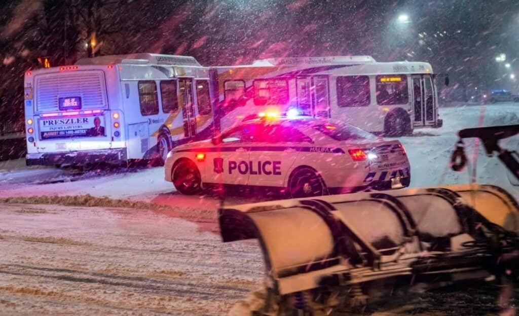 A snowy scene unfolds in Michigan, where two city buses and a police car with flashing lights navigate a snow-covered road alongside a snowplow, possibly due to a bus accident suit under investigation.