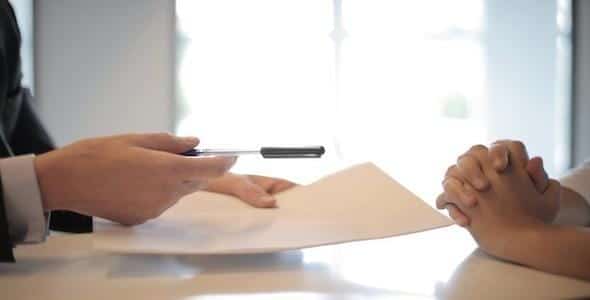 A person in a suit is handing over a settlement offer document and pen to another, whose hands are clasped on the table.