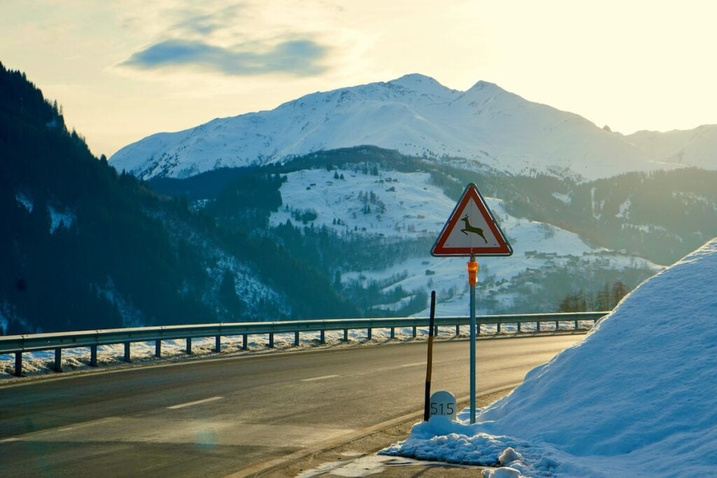 A winding mountain road with snow on the ground, a deer crossing warning sign, and distant snow-covered peaks under a cloudy sky—reminding drivers to stay alert and consult a Deer Accident Attorney if needed.