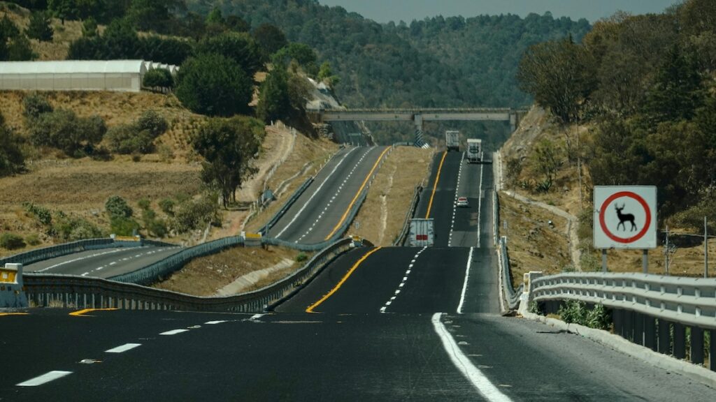 A highway with multiple lanes passes through hilly terrain. There are vehicles on the road and a deer-car accident lawyer warning sign visible on the right side, alerting drivers to potential wildlife crossings.