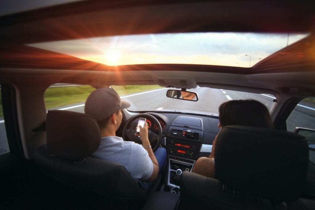 Two people sit in the front seats of a car driving on an empty road at sunset; the driver looks at a phone while steering, highlighting a scene where consulting a distracted driving lawyer might be necessary.